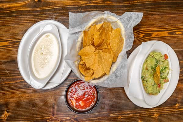 Basket of tortilla chips served with queso, guacamole, and chunky red salsa on a wooden table.