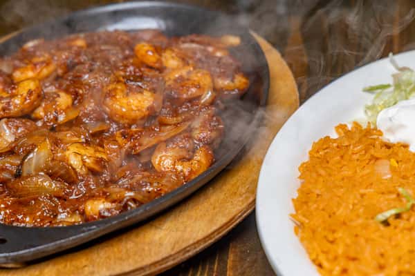 Sizzling shrimp and onions in a rich sauce served with Mexican rice, lettuce, and sour cream on a white plate.
