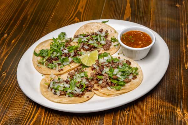 Street tacos with grilled beef, onions, and cilantro on corn tortillas, served with lime wedges and a side of red salsa.