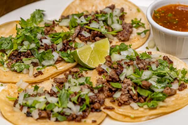 Street-style carne asada tacos topped with chopped onions and cilantro, served with lime wedges and a side of salsa.