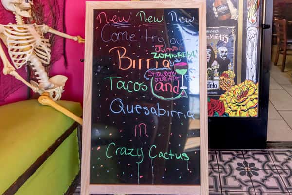 Colorful chalkboard sign promoting birria tacos, quesabirria, and strawberry cocktails at Crazy Cactus, next to a skeleton display.