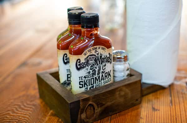 Two bottles of Gas Monkey Skidmark hot sauce are placed in a wooden caddy next to a salt shaker and paper towel roll on a wooden table.