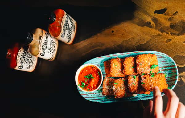 A hand reaches for a plate of crispy fried food with marinara sauce and parsley, alongside three bottles of Gas Monkey hot sauce.