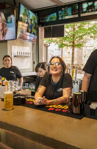 A smiling bartender with tattoos sits at the bar, surrounded by coworkers and a well-stocked tap system, with TVs and greenery visible in the background.