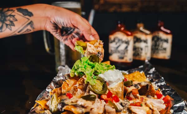 A hand adds guacamole to a plate of nachos topped with jalapeños, chicken, tomatoes, and sour cream, with bottles in the background.