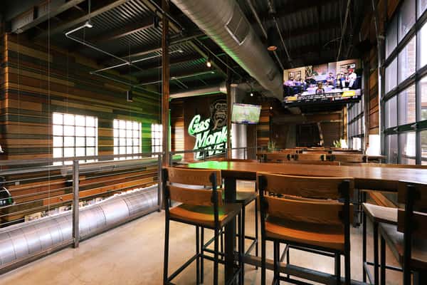 An interior view of the Gas Monkey Live! venue featuring high tables and chairs, with a view of a neon "Gas Monkey Live!" sign and two TVs displaying sports.