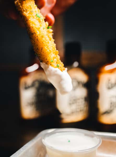 A hand dipping a fried item into a small cup of ranch sauce, with bottles of hot sauce visible in the background.