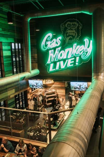 The interior of a lively venue with a "Gas Monkey Live!" neon sign, a merchandise area, and people dining and socializing below.