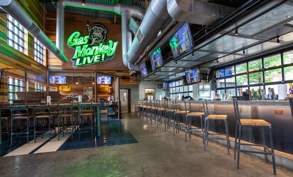 An interior view of Gas Monkey Live! featuring a neon "Gas Monkey Live!" sign, a bar with stools, and multiple TVs displaying content above.