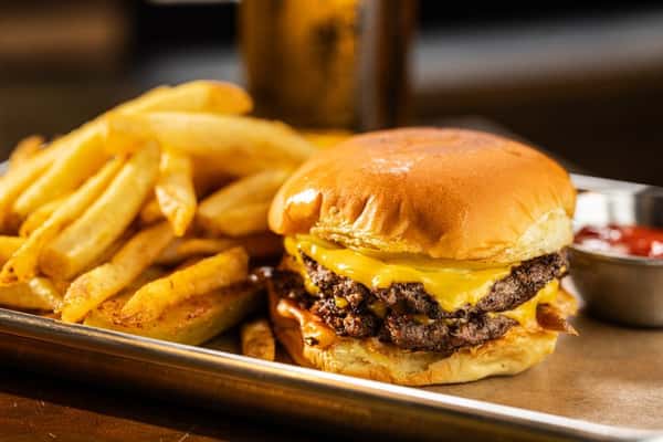 A close-up of a cheeseburger with two beef patties, melted cheddar cheese, served with crispy French fries and a side of ketchup.
