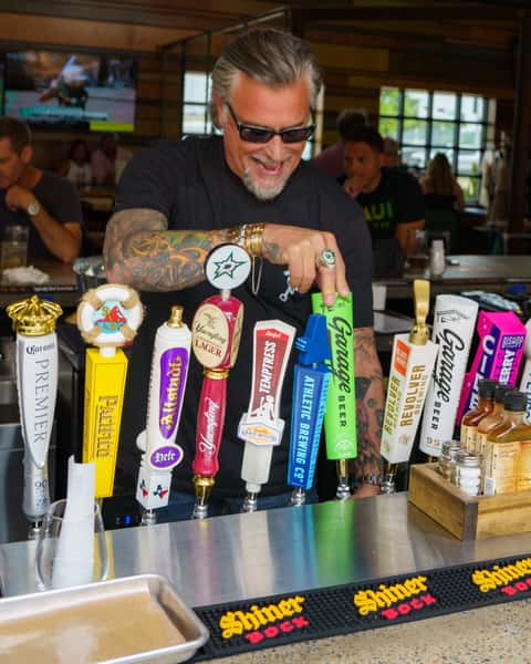 A man with tattoos and sunglasses is seen pouring a drink from a colorful selection of beer taps, including Garage Beer, Lakewood, and other brands.