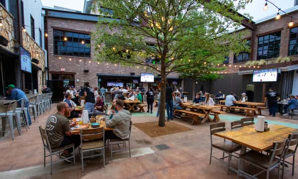 An outdoor patio area with string lights and several groups of people dining at tables, with TVs showing sports in the background.