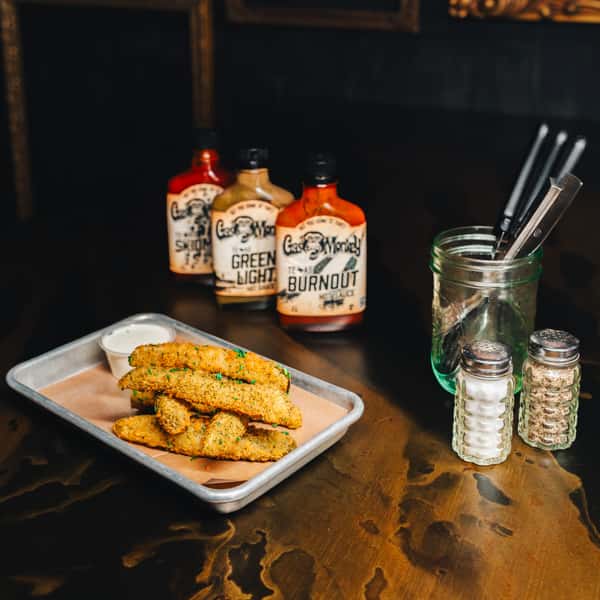 A tray of crispy fried pickles with a side of dipping sauce, alongside bottles of Gas Monkey hot sauce and salt and pepper shakers.
