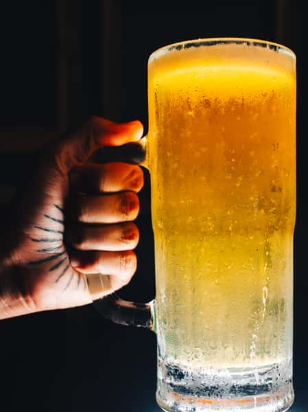 A hand grips a frosty mug of beer with condensation, the golden liquid glowing in low light.
