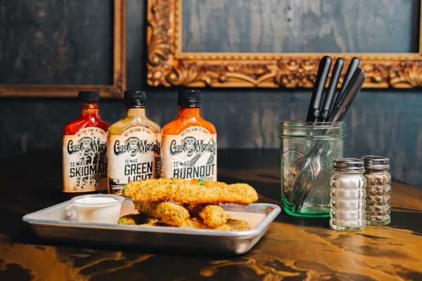 A tray of crispy fried food with dipping sauce sits next to three bottles of Gas Monkey hot sauce and a jar of utensils on a table.