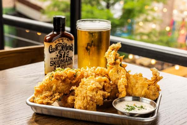 A tray of crispy fried chicken with a side of dipping sauce, accompanied by a cold glass of beer and a bottle of hot sauce.