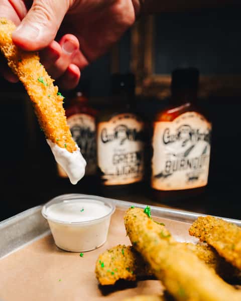 A hand dips a crispy fried pickle spear into ranch sauce, with bottles of hot sauce in the background.