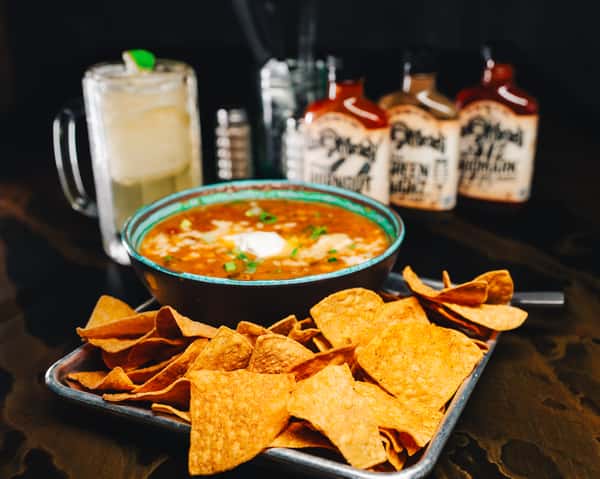 A bowl of chili with sour cream and green onions, accompanied by a side of tortilla chips. In the background, a margarita and bottles of hot sauce.