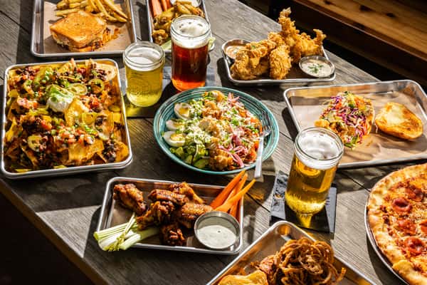 A spread of food and drinks, including nachos, fried chicken, a salad, wings with celery, a sandwich, pizza, and beers on the table.