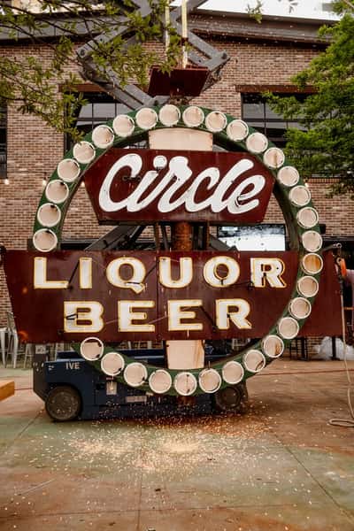 The "Circle Liquor Beer" sign is being worked on, with sparks flying as a worker on a lift repairs or installs it, surrounded by an outdoor seating area.
