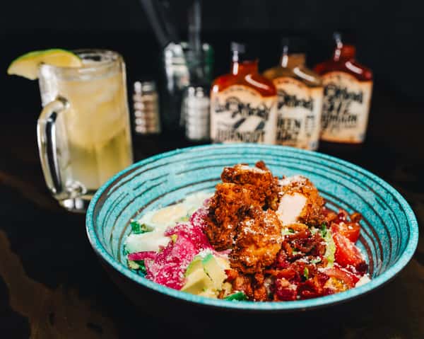 A vibrant salad with fried chicken, avocado, tomato, bacon, and cheese, served with a lime drink and hot sauce bottles in the background.