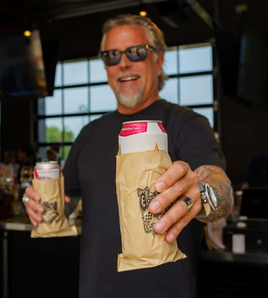 A man wearing sunglasses smiles while holding two cans of beer, each wrapped in a brown paper bag with a Gas Monkey logo.