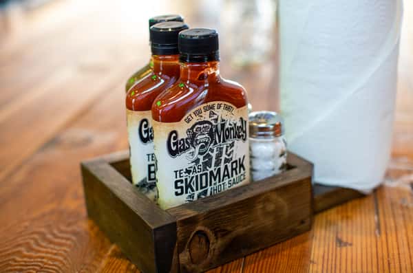 Two bottles of Gas Monkey Skidmark hot sauce are placed in a wooden caddy, with a salt shaker and paper towel roll in the background.