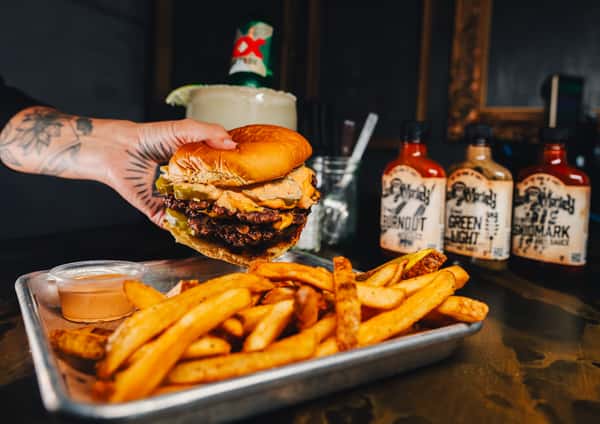 A hand holds a large burger with two patties, pickles, and sauce, accompanied by a side of fries and bottles of hot sauce.