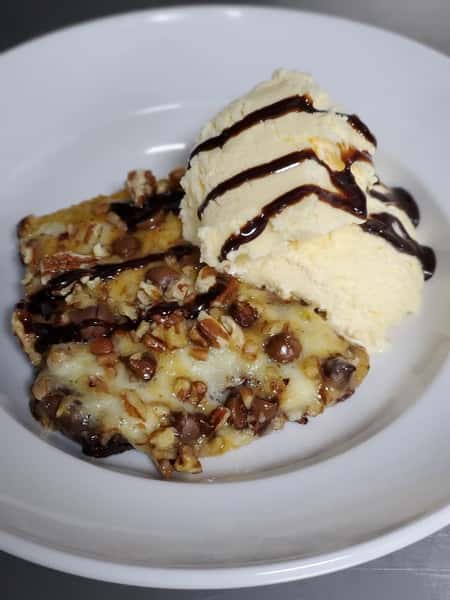 A slice of cake topped with nuts, chocolate syrup, and a scoop of vanilla ice cream