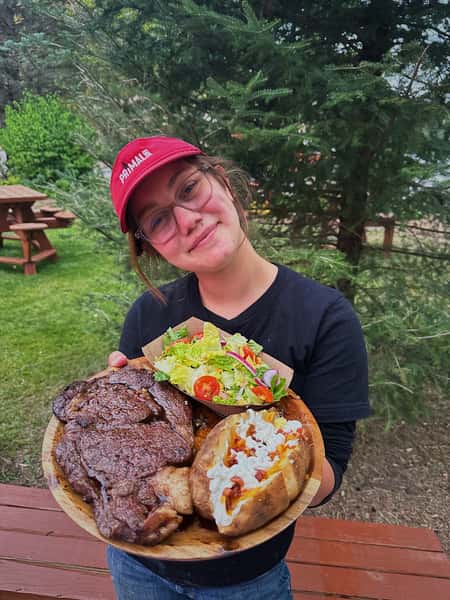 A brunette woman wearing glasses and a red baseball cap that says "Primal Eats" holding up a ribeye platter with a baked potato and side salad.