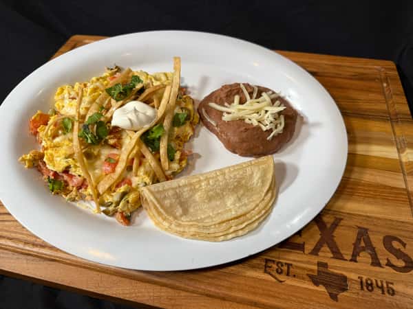 Breakfast plate with scrambled eggs, tortilla strips, refried beans topped with cheese, and folded corn tortillas.