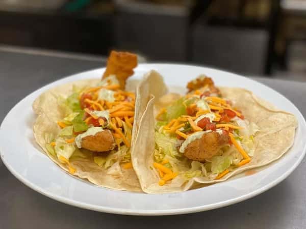 Two fish tacos with shredded lettuce, cheddar cheese, diced tomatoes, and creamy sauce in soft flour tortillas.