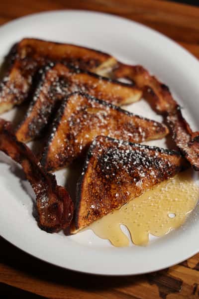 Golden brown French toast dusted with powdered sugar, served with crispy bacon strips and maple syrup on a white plate.