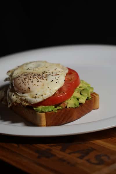 Avocado toast topped with sliced tomato and a fried egg, seasoned with black pepper, served on a white plate.
