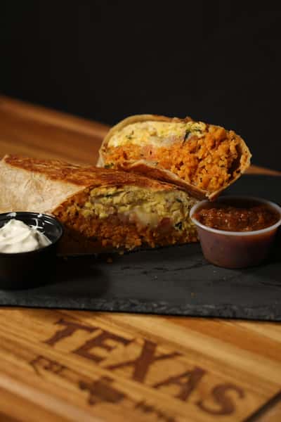 Grilled breakfast burrito cut in half, filled with rice and eggs, served with sour cream and salsa on a wooden tray.
