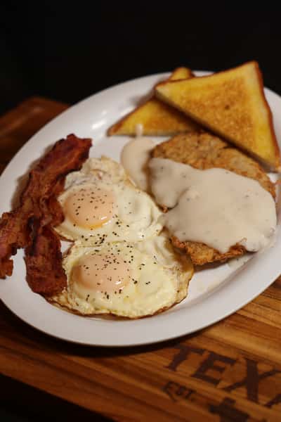 Country-style breakfast plate with two fried eggs, crispy bacon strips, toast, and chicken fried steak topped with creamy gravy