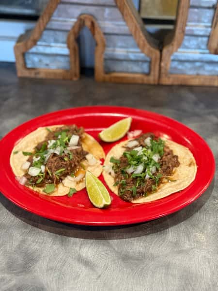 Two beef tacos garnished with chopped onions and cilantro, served with lime wedges on a red plate.