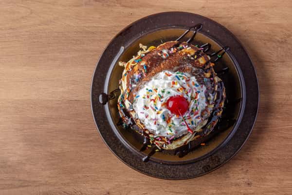 Stack of pancakes topped with whipped cream, sprinkles, chocolate drizzle, and cherry, indulgent diner dessert