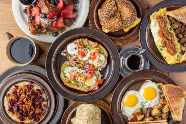 Assorted American brunch spread with French toast, bagel sandwich, omelet, avocado toast, eggs, and coffee on rustic table at One Oakland