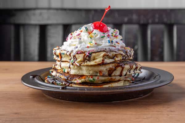 Stack of pancakes topped with whipped cream, rainbow sprinkles, chocolate drizzle, and cherry, indulgent diner dessert