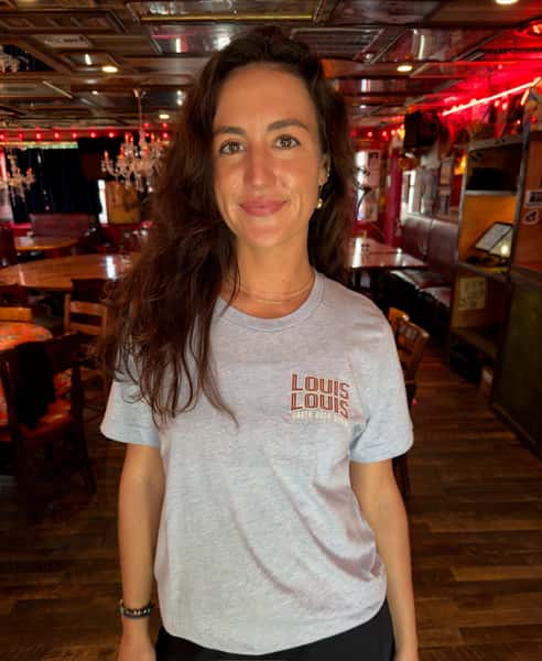 a woman wearing a "LOUIS LOUIS" branded gray t-shirt, standing inside a restaurant with red lighting and wooden tables in the background.