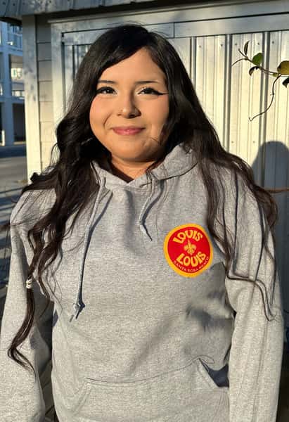 A woman wearing a gray hoodie with a "LOUIS LOUIS" logo stands outside, smiling in front of a metal structure.