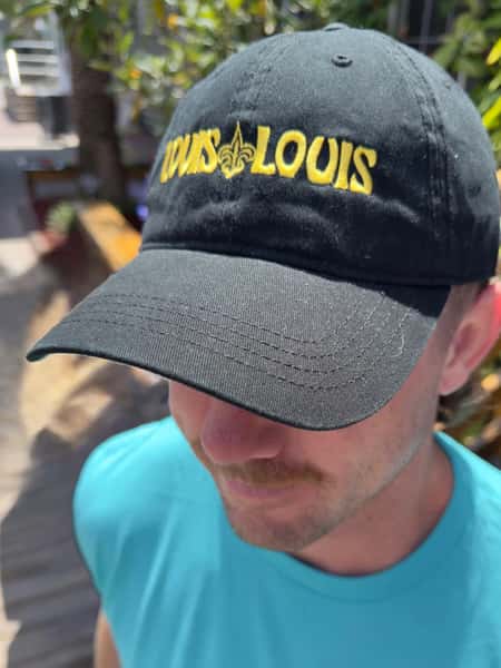 A black baseball cap featuring the "LOUIS LOUIS" logo in yellow with a fleur-de-lis design in between the words.