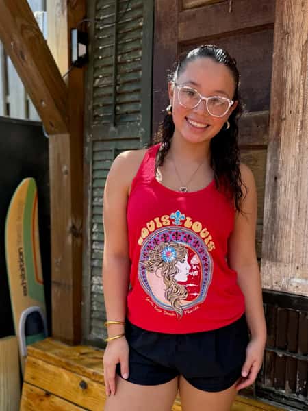 A woman smiles while standing outside in a red tank top with a colorful "Louis Louis" graphic, surrounded by wooden structures and a surfboard.