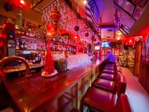 A vibrant bar area decorated with red lighting, hanging ornaments, and a festive atmosphere, showing a well-stocked bar and stools.