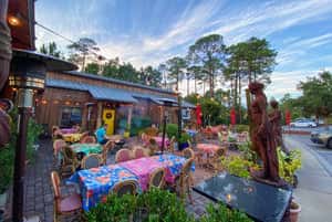 An outdoor dining area with colorful tables, plants, and a statue, featuring a relaxed atmosphere and rustic decor under a beautiful sky.