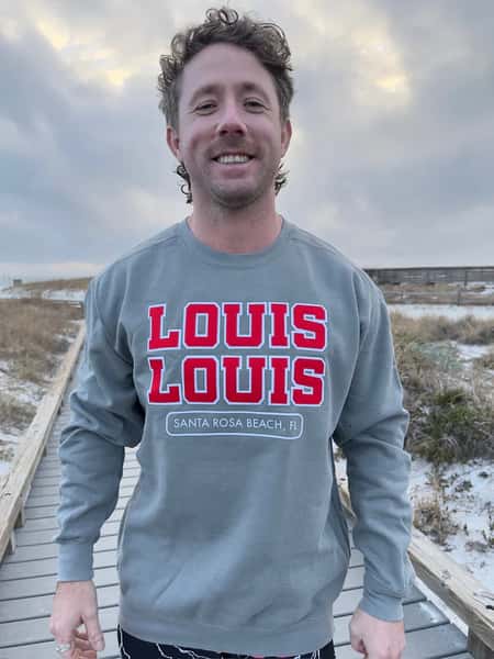 A person wearing a "LOUIS LOUIS" sweatshirt while standing outdoors on a boardwalk with a cloudy sky in the background.