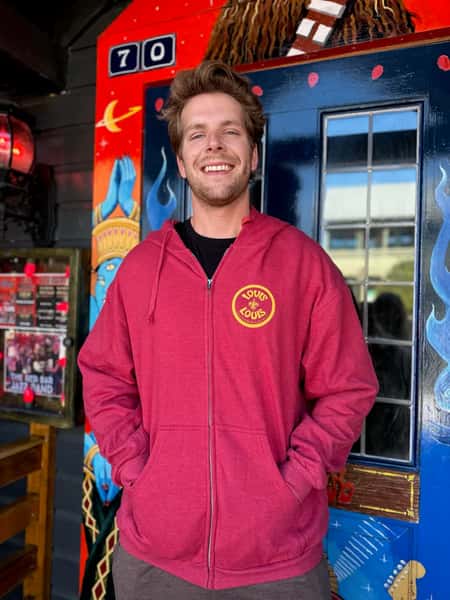 A person standing outdoors, smiling, wearing a red zip-up hoodie with the "LOUIS LOUIS" logo on it. The background features a colorful mural.