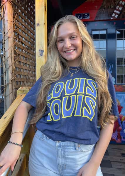 A woman wearing a blue "LOUIS LOUIS" t-shirt with yellow lettering. She is standing outside in front of a wooden structure with painted artwork behind her.