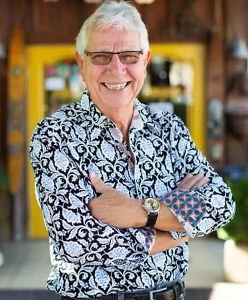 A man in a black and white paisley shirt smiles while standing with his arms crossed outside a restaurant.